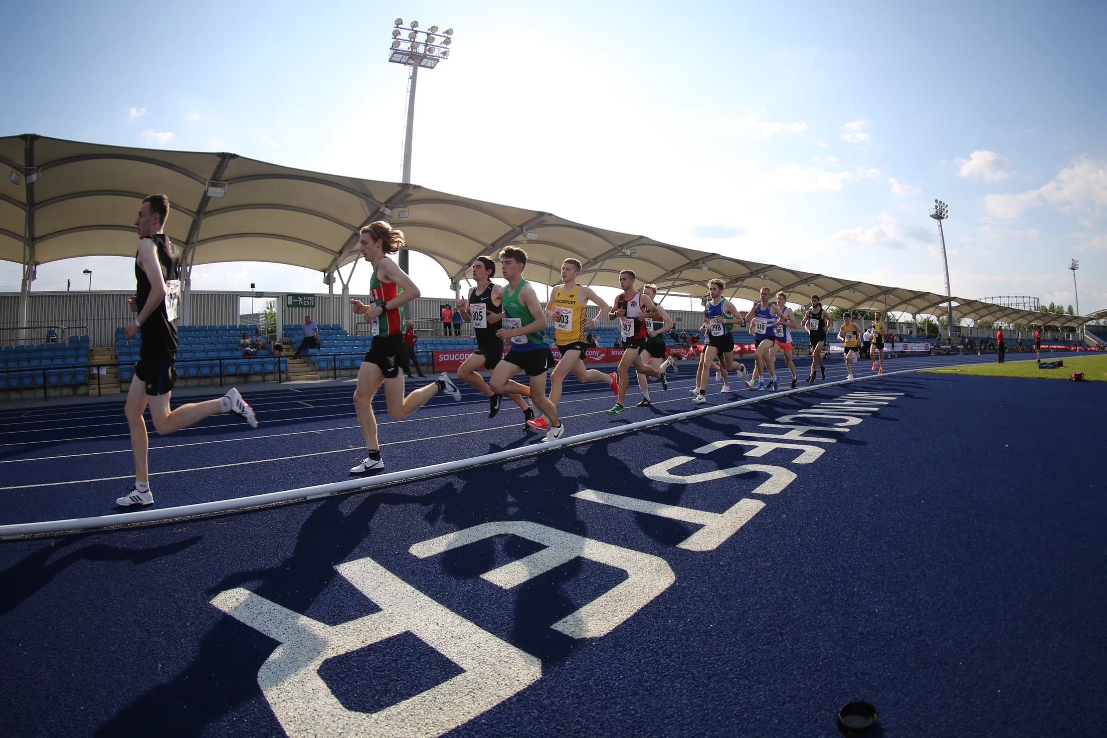 Pack of club runners mid-race on the blue track at Manchester